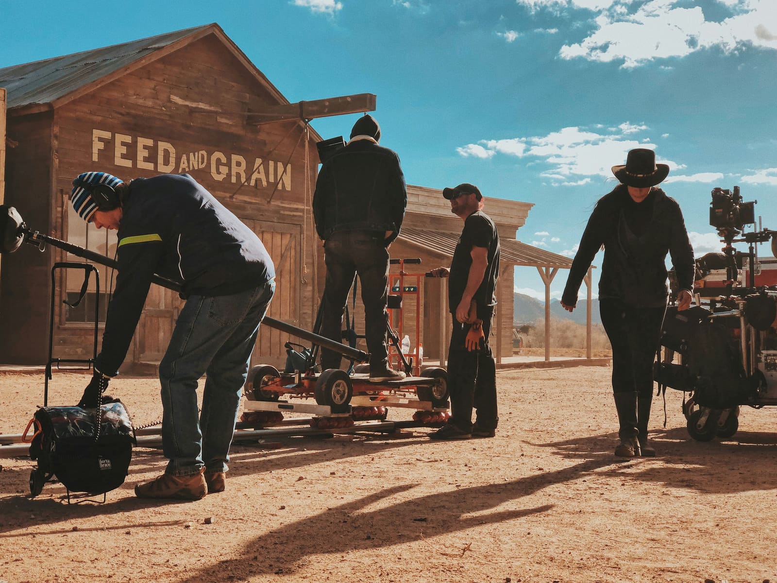 A group of filmmakers on a western film set, surrounded by rustic buildings and desert scenery, capturing a scene with a camera and crew.