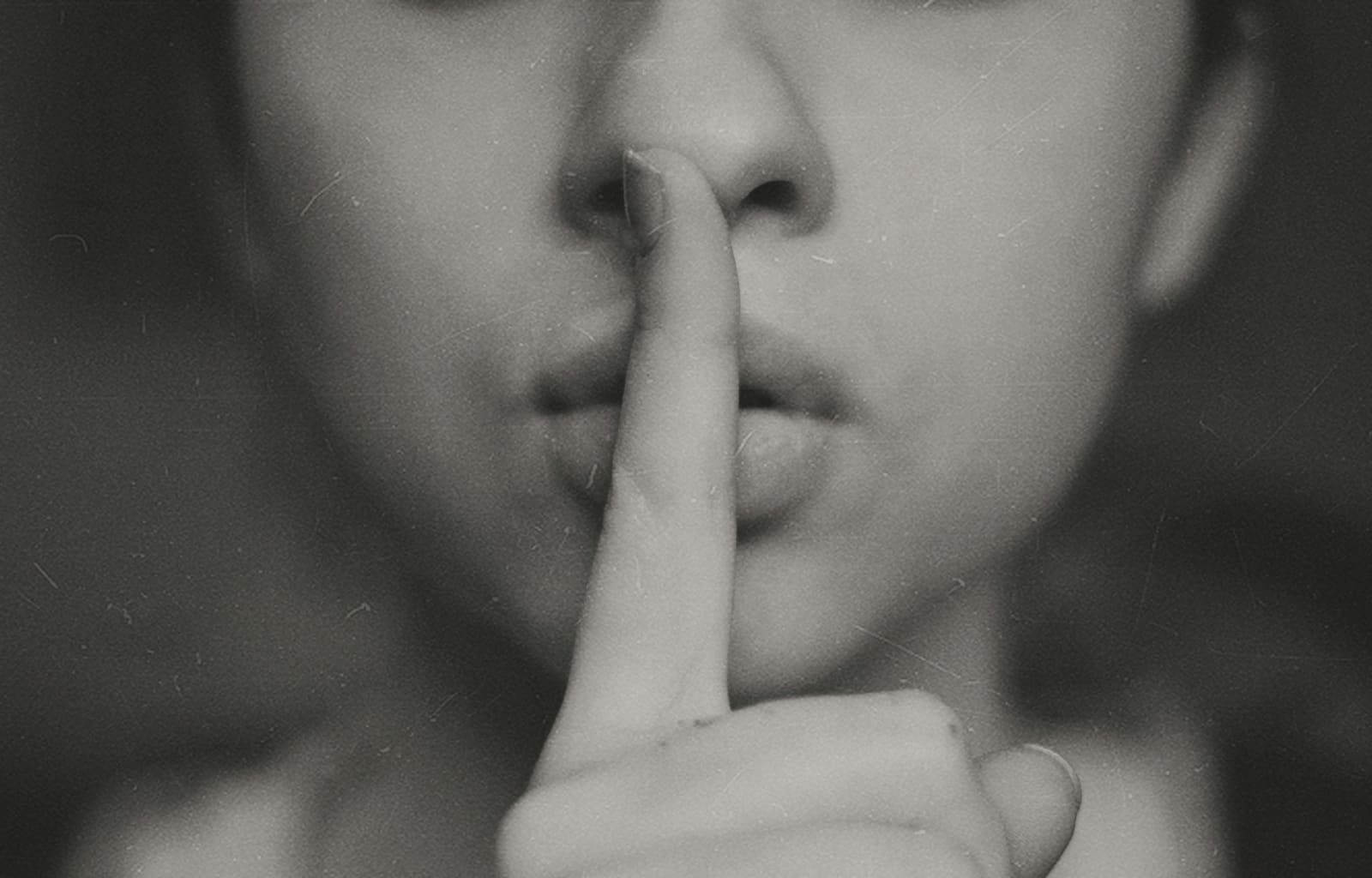 A film director holds a finger to their lips, signalling for silence on a busy film set during the recording of a silent scene. Crew members freeze and comply, with cameras and lighting equipment visible in the background.